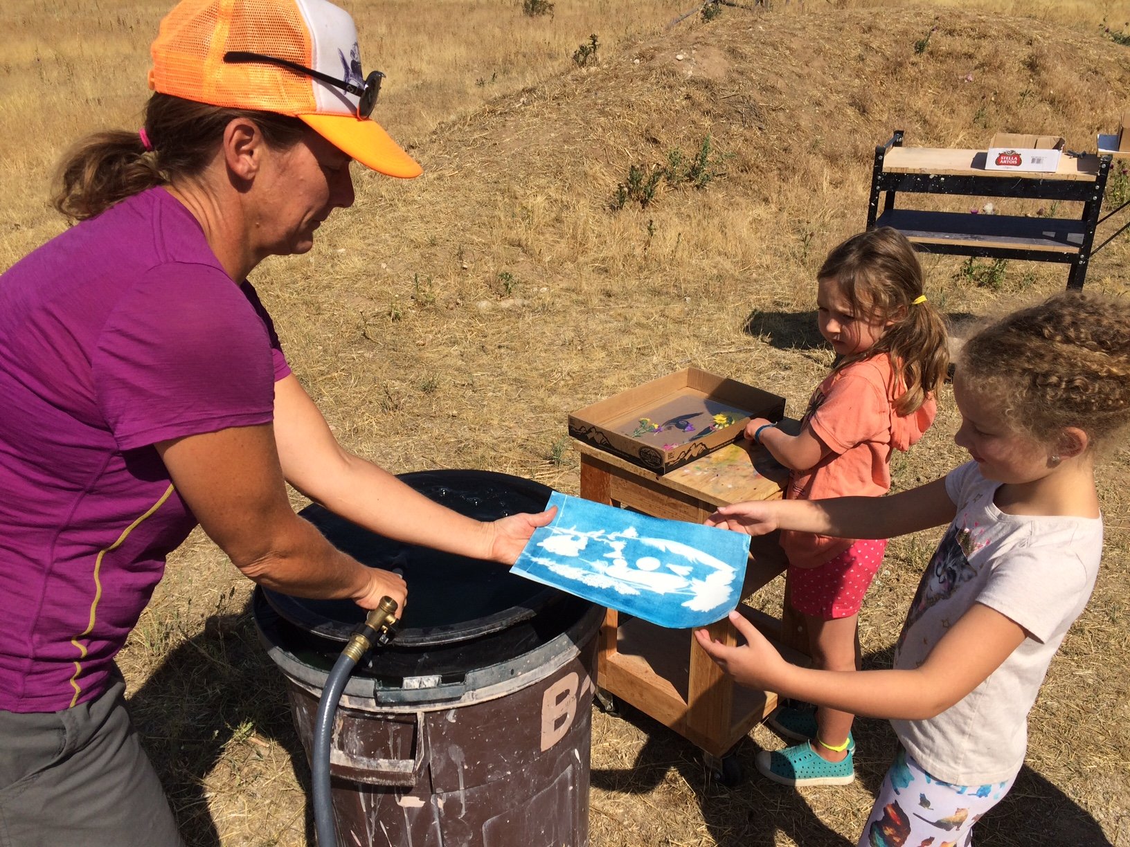 Teton Valley Non-Profits, An energetic adult assists two eager children at an outdoor activity station, gently rinsing a blue piece of paper while standing on a dry, grassy hill. This heartwarming scene exemplifies the type of community engagement that the Community Foundation of Teton Valley facilitates through its philanthropic efforts. By channeling charitable donations and support to local non-profits and businesses, the Foundation ensures enriching educational experiences such as this one are made possible for individuals of all ages in our community. The Tin Cup Challenge & Run is the CFTV annual giving event day.
