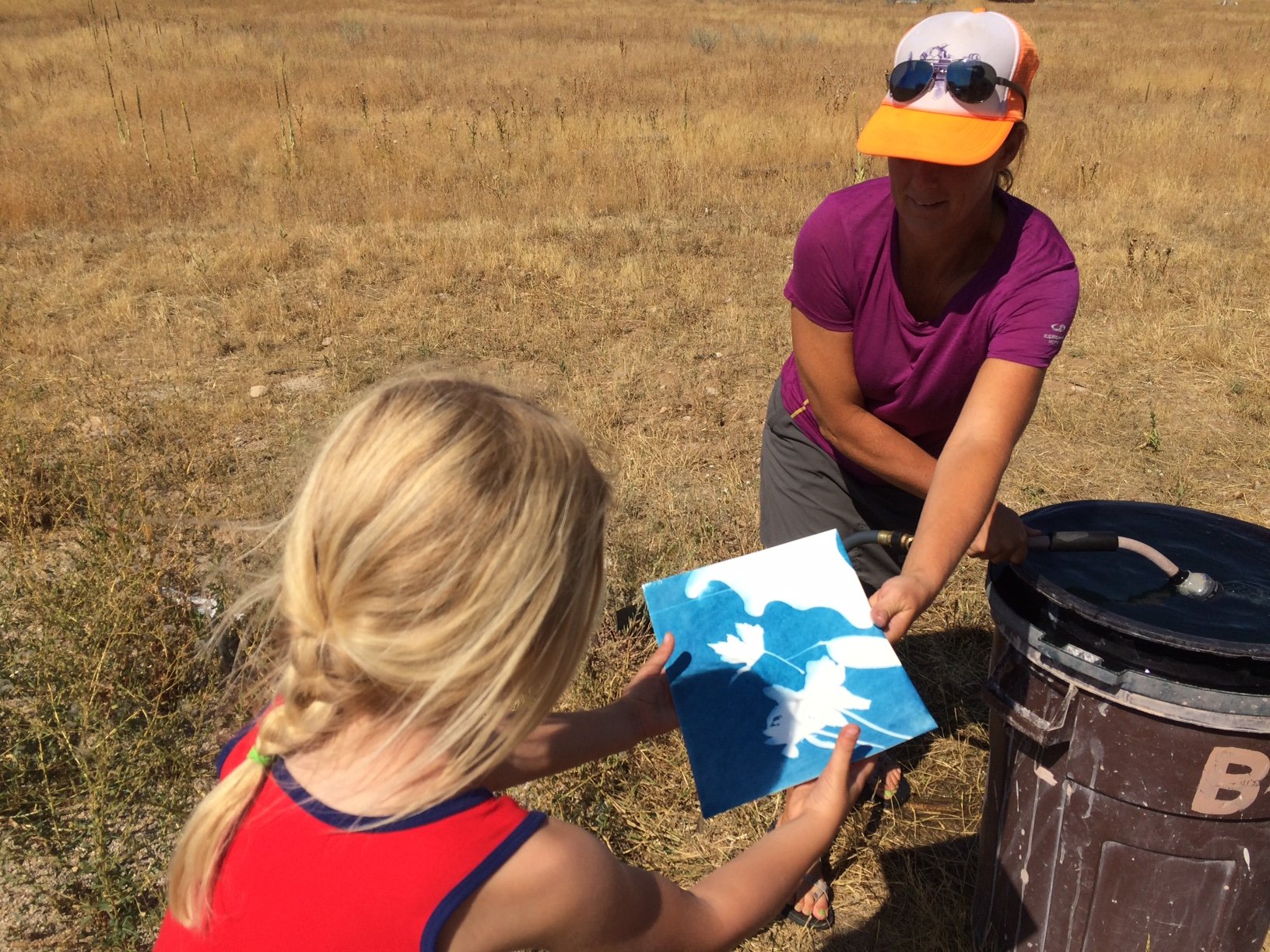 Teton Valley Non-Profits, In a sunlit open space adorned with dry grasses, an adult in a purple shirt and orange cap gently hands a blue and white art piece to a child with braided hair. This simple yet poignant moment captures the essence of how the Community Foundation of Teton Valley empowers and uplifts the community. By fostering charitable giving, this organization not only supports local non-profits but also strengthens businesses, creating an environment where culture, art, education, and human connection can thrive. The transfer of this artwork symbolizes the ripple effect of generosity—from donors to foundations to recipients—mirroring how collective efforts enrich lives in Teton Valley. The Tin Cup Challenge & Run is the CFTV annual giving event day.