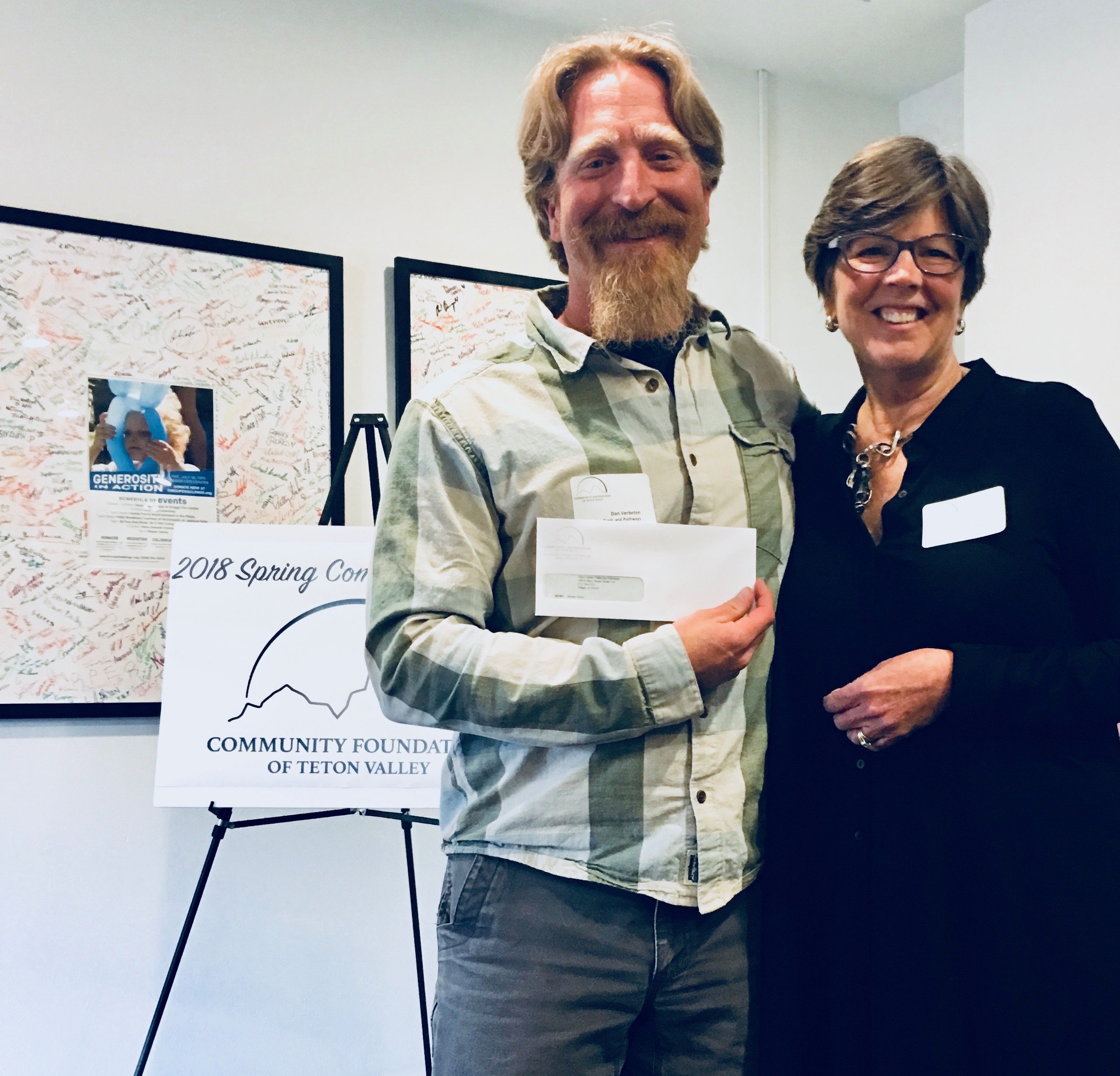 Teton Valley Non-Profits, A man and a woman stand together indoors, beaming with pride and joy. The man holds an envelope, which likely contains a donation or grant. Behind them, a sign reads "2018 Spring Comm. Community Foundation of Teton Valley," highlighting the event organized by the Community Foundation of Teton Valley. This foundation plays a crucial role in supporting local non-profits and businesses through charitable giving, fostering community growth and sustainability by providing necessary resources to those dedicated to making a positive impact in the region. The Tin Cup Challenge & Run is the CFTV annual giving event day.